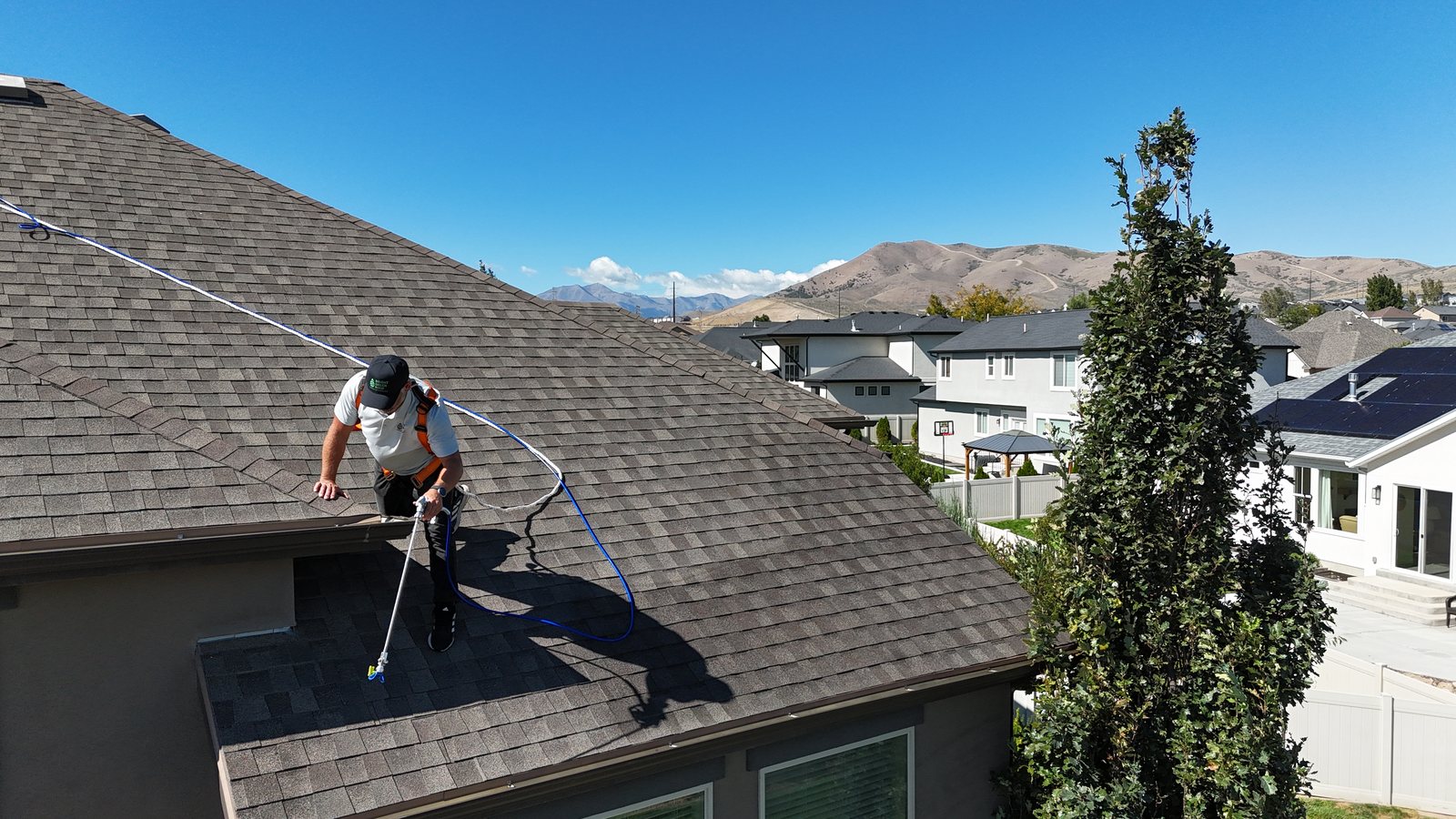 An applicator in a fall-protection harness kneeling at the edge of a Saratoga Springs roof, spraying the shingles with a wand, mountains behind.