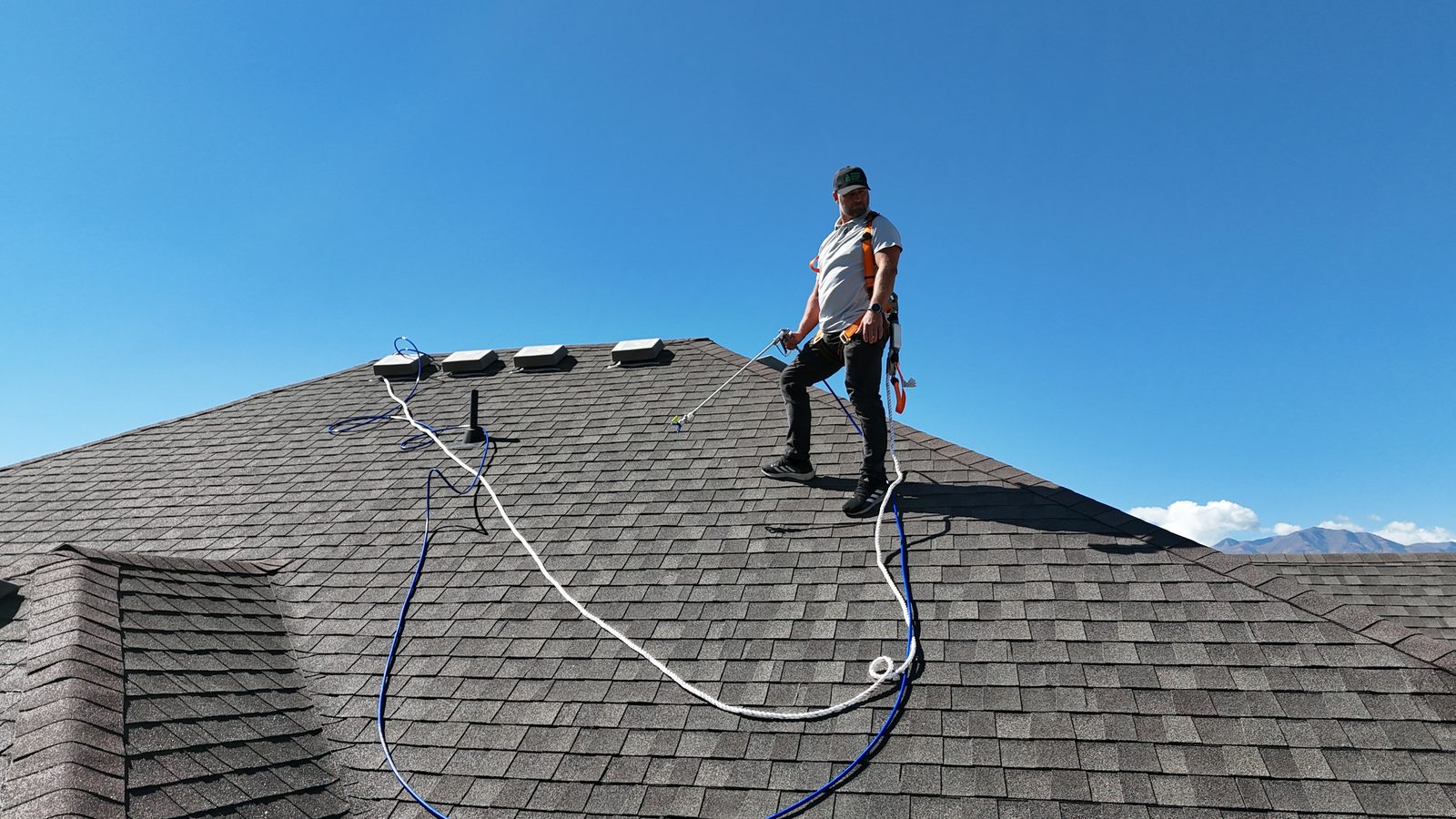 An applicator standing near the ridge in a fall-protection harness, rope and spray hose running down the roof.